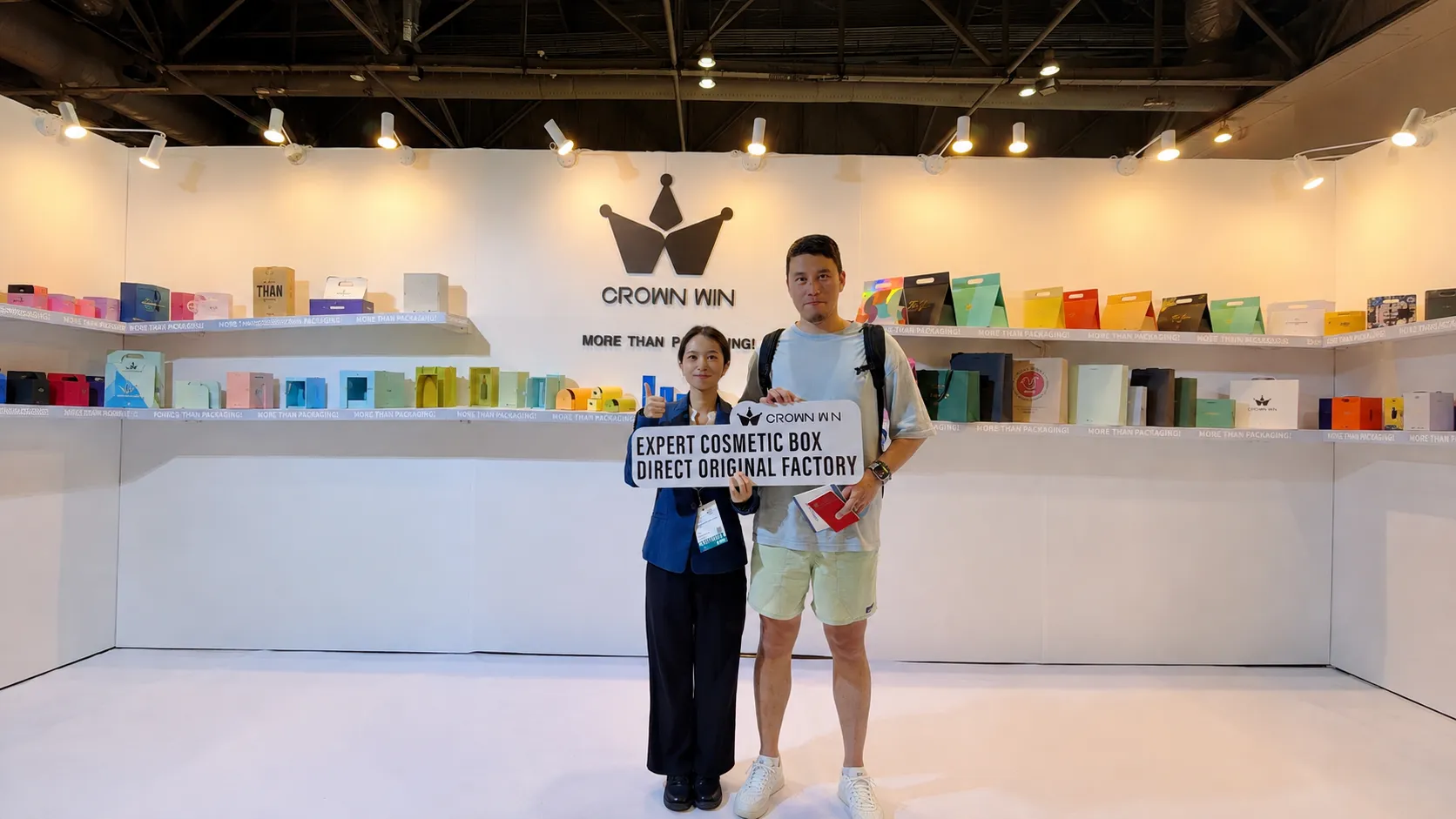 Crown Win staff and male trade visitor hold factory branding sign at Hong Kong packaging fair, full range custom paper packaging on display